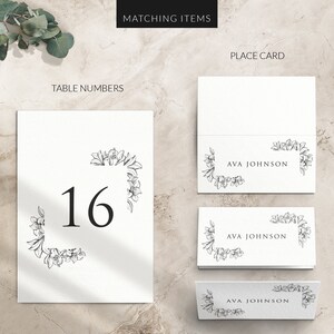 May include: White wedding table number card with a black floral border and the number 16 in black font. Matching place cards with the name "AVA JOHNSON" in black font are also shown.