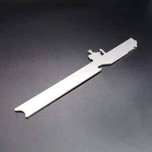 Op de afbeelding: Een zilveren metalen flessenopener in de vorm van een silhouet van een vliegdekschip. De flessenopener heeft een uitsparing om flessen te openen.