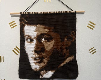 May include: A brown crocheted wall hanging featuring a portrait of a person. The hanging is attached to a wooden dowel rod and is decorated with gold accents.