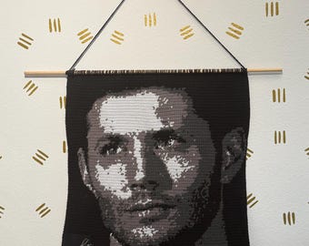 May include: A black, gray, and white crocheted portrait of a man, hanging from a wooden dowel and black string. The portrait is signed in the lower left corner. The background is a white wall with gold dashes.