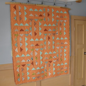 May include: A quilt with a geometric pattern of orange and teal triangles. The quilt is hanging on a wall.