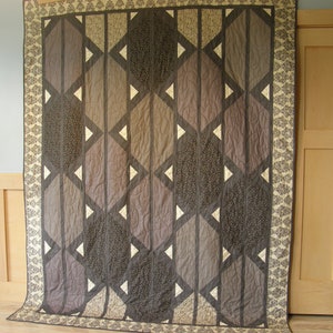May include: A large quilt with a geometric pattern in shades of brown and cream. The quilt is hanging on a wall and has a decorative border.