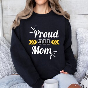 以下が含まれることがあります： 黒のトレーナーに「Proud 911 Mom」という白い文字と黄色の文字が書かれています。文字の周りは黄色の矢印と星で囲まれています。