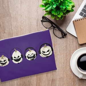 May include: Purple zippered pouch with a white background and five black and white illustrations of pumpkins wearing headphones. The pumpkins have different facial expressions.