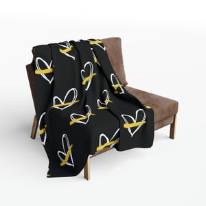 Puede incluir: Manta de tiro negra con un patrón de corazón blanco y detalles dorados. La manta está drapeada sobre un sillón marrón.
