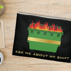 Puede incluir: Una bolsa negra con cremallera con un contenedor de basura verde en llamas y el texto "ASK ME ABOUT MY SHIFT".