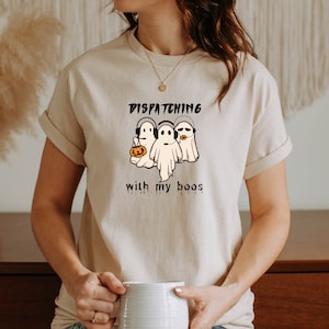 Puede incluir: Camiseta beige con un diseño de Halloween. El diseño presenta tres fantasmas de dibujos animados con auriculares, con el texto "Dispatching with my boos". Un fantasma sostiene una calabaza.