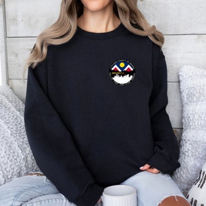 May include: Black sweatshirt with a circular graphic featuring the Denver, Colorado city flag. The flag depicts a yellow sun, blue mountains, and a white cityscape with the text "City and County of Denver" and "Mile High City" around the graphic.
