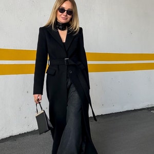 May include: A woman wearing a black coat with a belt, black pants, and black sunglasses. She is holding a small black handbag.