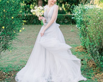 May include: A woman in a flowing, light gray wedding dress with a deep V-neck and a bouquet of white flowers. The dress has a tulle skirt and a bow in the back. The background is a garden with green foliage.