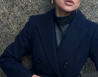 Puede incluir: Un abrigo azul marino de doble botonadura con una parte superior negra y un bolso de mano negro. El bolso de mano tiene el texto "SAINT LAURENT" impreso. La joyería de oro incluye una pulsera, anillos y pendientes. También se ve un reloj de pulsera con esfera dorada.