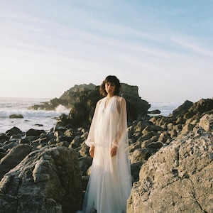 May include: A woman wearing a white, sheer, long-sleeved dress stands on a rocky beach. The dress has a V-neckline and a flowing skirt. The woman is looking at the camera.
