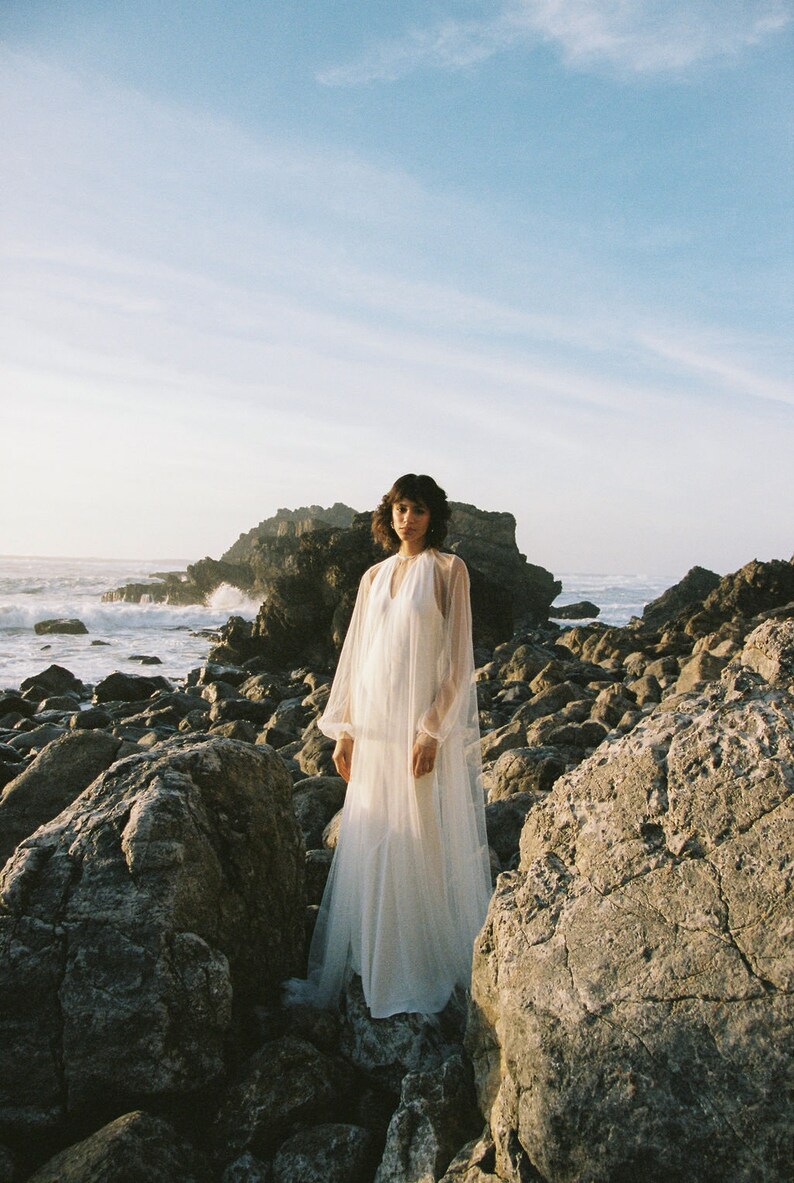 May include: A woman wearing a white, sheer, long-sleeved dress stands on a rocky beach. The dress has a V-neckline and a flowing skirt. The woman is looking at the camera.