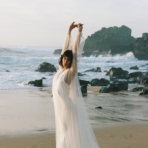 May include: A woman wearing a white wedding dress with a long veil, standing on a sandy beach with the ocean in the background.