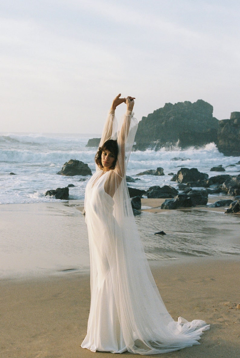 May include: A woman wearing a white wedding dress with a long veil, standing on a sandy beach with the ocean in the background.
