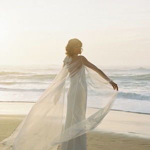 May include: A woman wearing a white wedding dress with a sheer cape flowing in the wind on a beach.