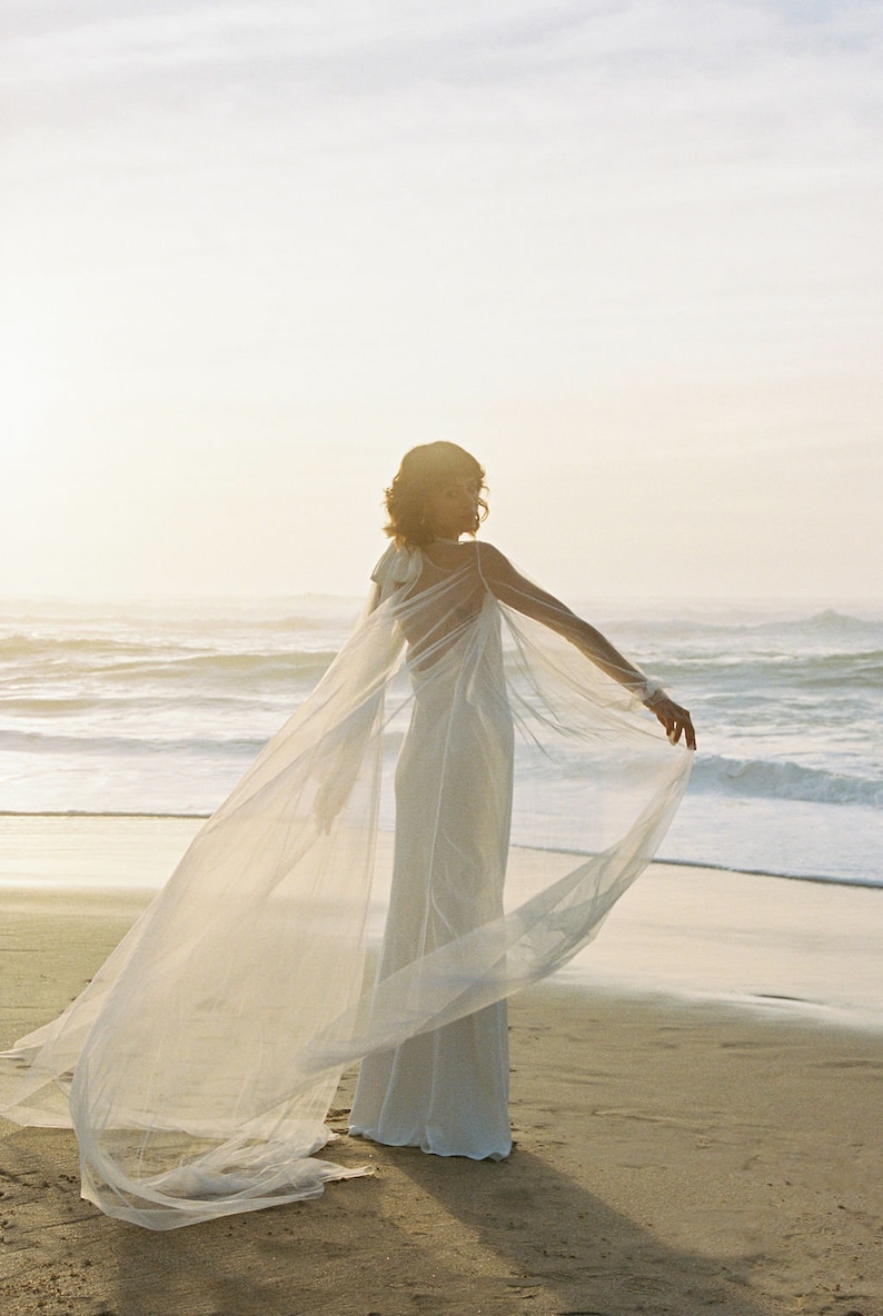 May include: A woman wearing a white wedding dress with a sheer cape flowing in the wind on a beach.
