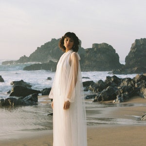 May include: A woman wearing a white sheer gown with long sleeves and a high neckline stands on a sandy beach with rocky cliffs in the background.