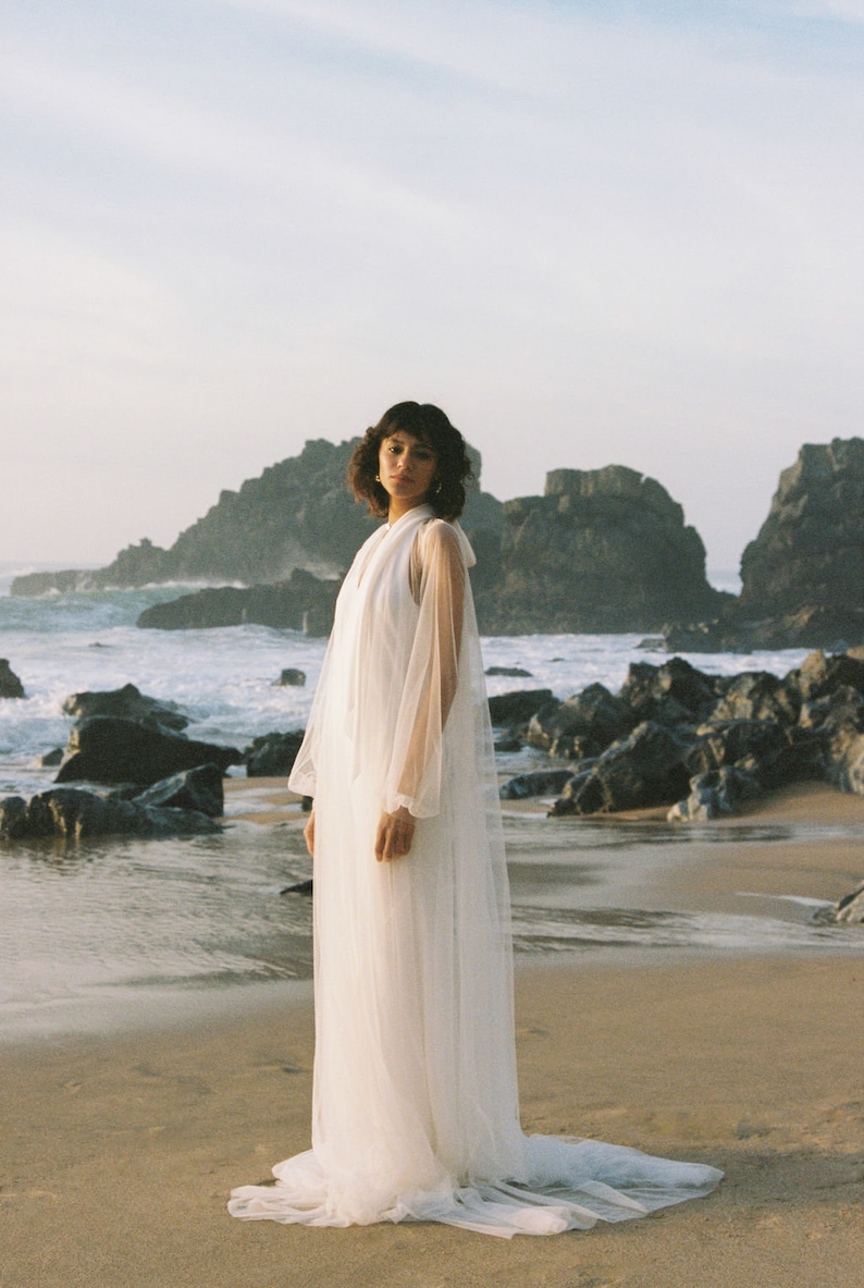 May include: A woman wearing a white sheer gown with long sleeves and a high neckline stands on a sandy beach with rocky cliffs in the background.