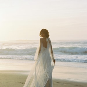 May include: A woman in a white wedding dress with a long veil walks on a sandy beach with the ocean in the background.