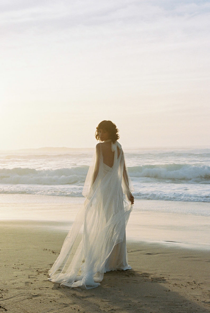 May include: A woman in a white wedding dress with a long veil walks on a sandy beach with the ocean in the background.