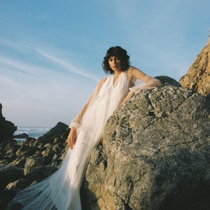 May include: A woman wearing a white sheer dress with long sleeves poses on a large rock by the ocean. The dress has a flowing skirt and a high neckline.