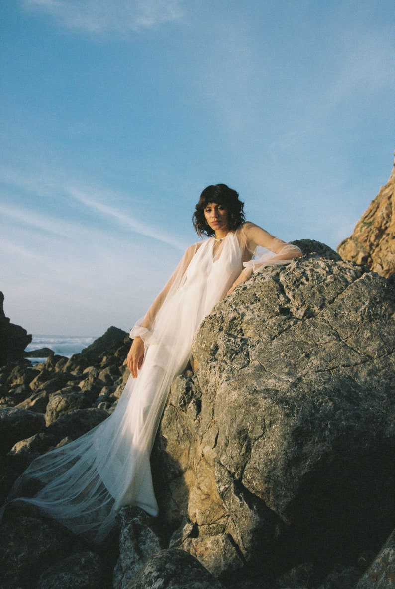 May include: A woman wearing a white sheer dress with long sleeves poses on a large rock by the ocean. The dress has a flowing skirt and a high neckline.