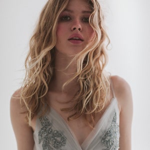May include: A woman with long, wavy blonde hair wearing a light gray dress with silver beaded embellishments. The dress has a V-neck and thin straps. The background is a plain white.