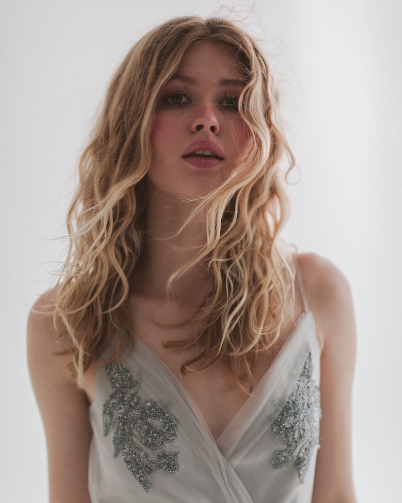 May include: A woman with long, wavy blonde hair wearing a light gray dress with silver beaded embellishments. The dress has a V-neck and thin straps. The background is a plain white.