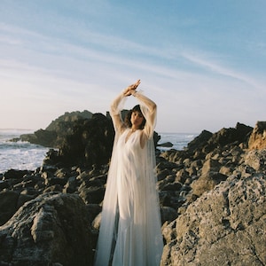 May include: A woman wearing a white gown with a sheer overlay stands on a rocky beach. The gown has long sleeves and a V-neckline. The woman is looking at the camera with her arms raised above her head.