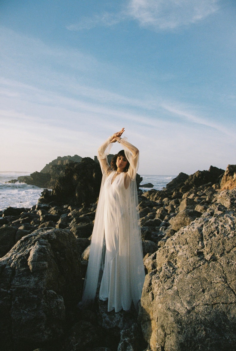 May include: A woman wearing a white gown with a sheer overlay stands on a rocky beach. The gown has long sleeves and a V-neckline. The woman is looking at the camera with her arms raised above her head.