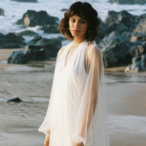 May include: A woman wearing a white sheer dress with long sleeves and a high neckline. She is standing on a beach with rocks and water in the background.