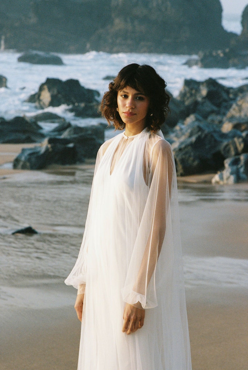 May include: A woman wearing a white sheer dress with long sleeves and a high neckline. She is standing on a beach with rocks and water in the background.
