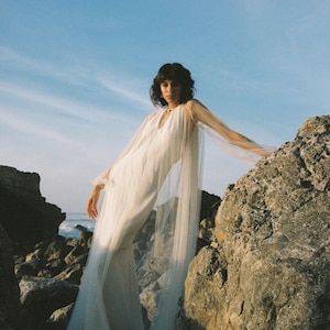 May include: A woman wearing a white dress with a sheer overlay stands on a rocky cliff. The dress has a V-neckline and long sleeves. The woman is looking at the camera.