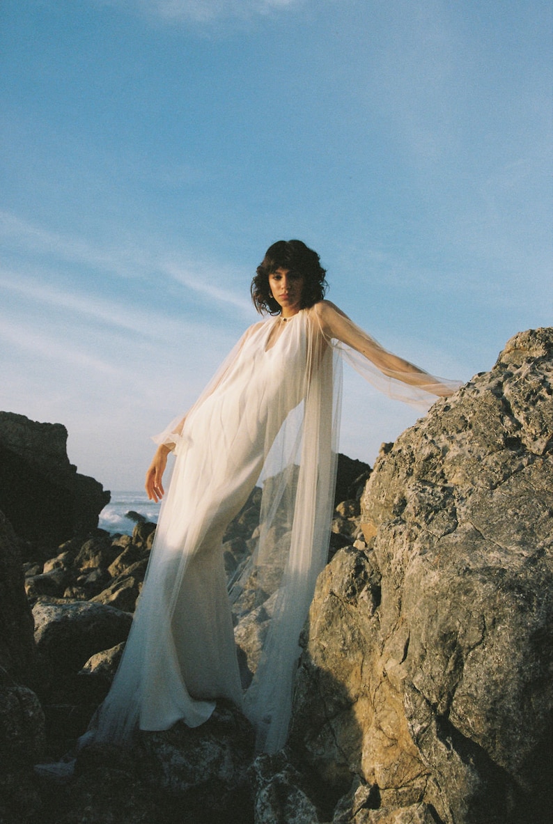 May include: A woman wearing a white dress with a sheer overlay stands on a rocky cliff. The dress has a V-neckline and long sleeves. The woman is looking at the camera.