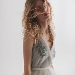 May include: A light gray dress with a V-neck bodice adorned with silver beading. The dress features a white tulle skirt and a white belt. The model has long, wavy blonde hair.