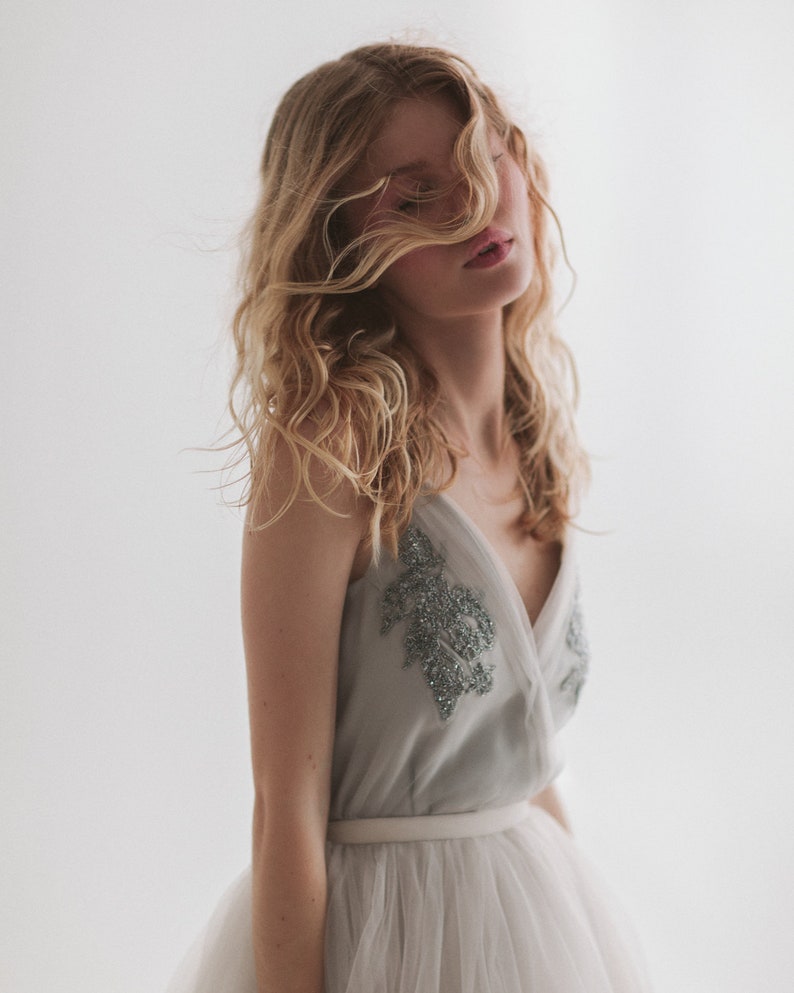 May include: A light gray dress with a V-neck bodice adorned with silver beading. The dress features a white tulle skirt and a white belt. The model has long, wavy blonde hair.