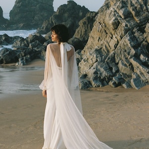 May include: A woman wearing a white wedding dress with a long tulle veil, standing on a sandy beach with rocky cliffs in the background.