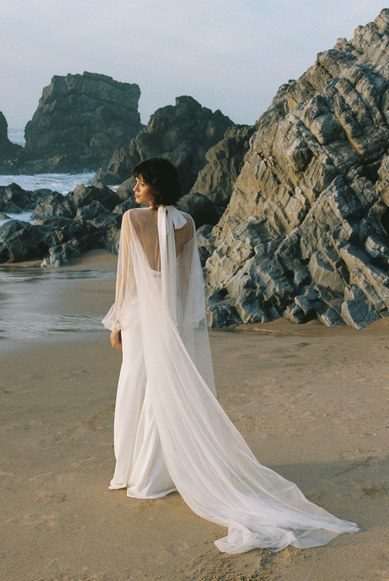 May include: A woman wearing a white wedding dress with a long tulle veil, standing on a sandy beach with rocky cliffs in the background.