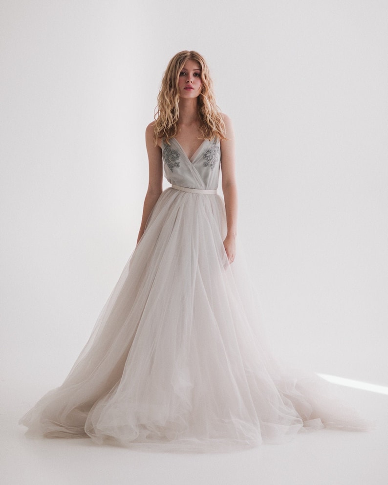 May include: A light gray wedding dress with a tulle skirt and a bodice with silver embroidery. The dress has a V-neckline and a thin, light-colored belt. The skirt flows to the floor.