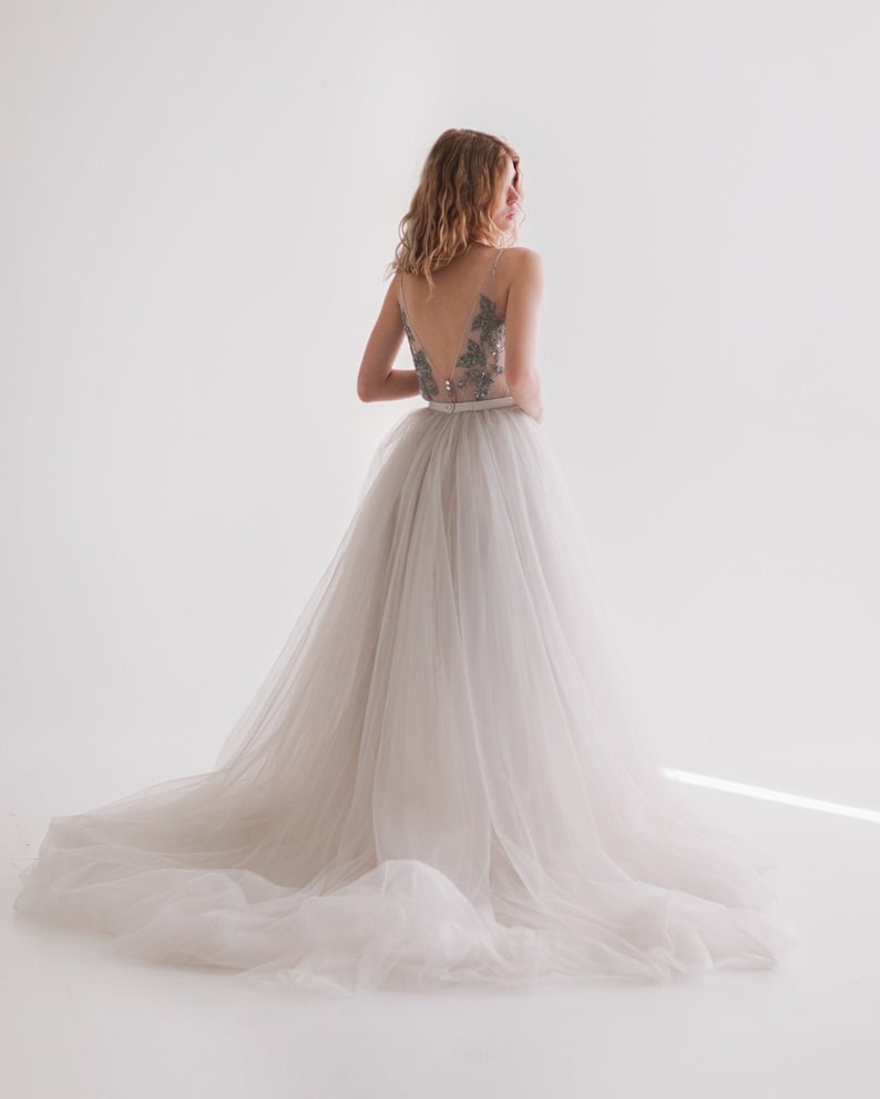 May include: A light-colored wedding dress with a tulle skirt and a beaded bodice. The dress has a V-neckline and a long train. The bride is turned away from the camera, with her back to the viewer.