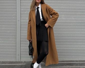 May include: A woman wearing a brown coat, a white collared shirt with a black tie, a black skirt, black tights, and black shoes. She is standing in front of a gray wall.