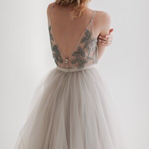 May include: A light gray tulle skirt paired with a sheer top adorned with silver embellishments. The top features a low V-neck and delicate straps. A thin belt cinches the waist, creating an elegant silhouette. The dress is ideal for special occasions.