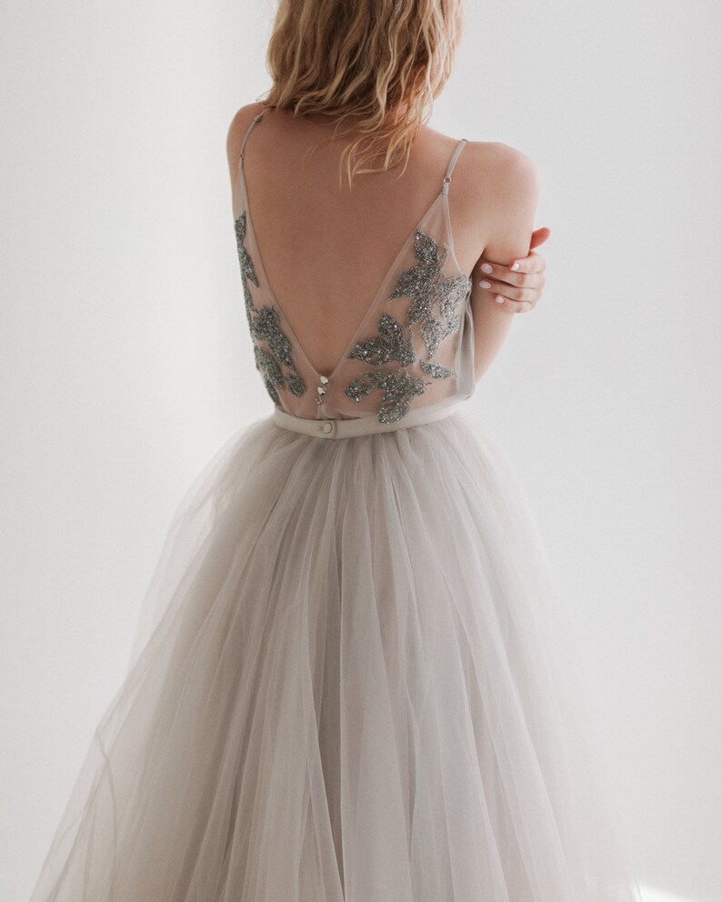 May include: A light gray tulle skirt paired with a sheer top adorned with silver embellishments. The top features a low V-neck and delicate straps. A thin belt cinches the waist, creating an elegant silhouette. The dress is ideal for special occasions.