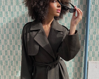 Puede incluir: Una mujer con un abrigo verde oscuro con cinturón y un cuello grande. Está sosteniendo un par de gafas de sol negras en su mano derecha.