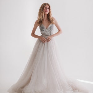 May include: A light gray wedding dress with a tulle skirt and a beaded bodice. The dress has a V-neckline and a flowing, floor-length skirt. The model is standing in front of a white background.
