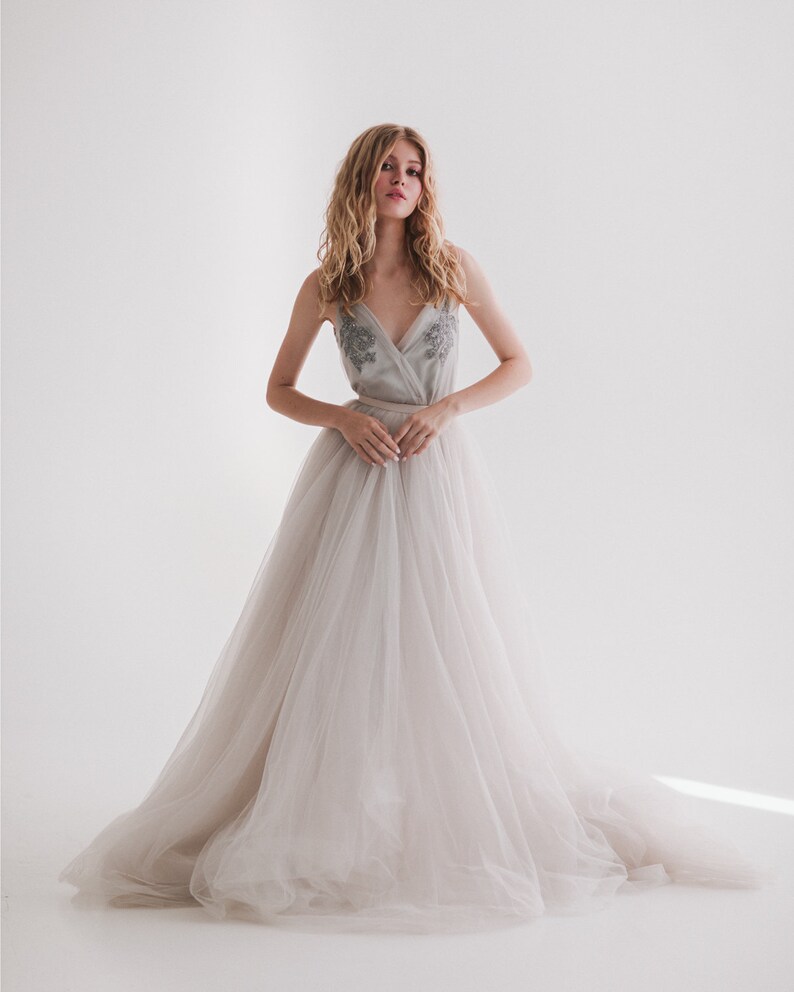May include: A light gray wedding dress with a tulle skirt and a beaded bodice. The dress has a V-neckline and a flowing, floor-length skirt. The model is standing in front of a white background.