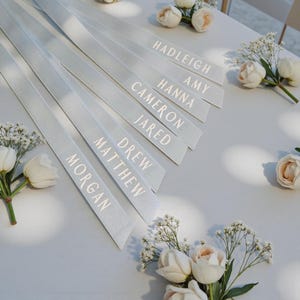 May include: A set of light grey satin ribbons, each printed with a white name including Hadleigh, Amy, Hanna, Cameron, Jared, Drew, Matthew, and Morgan. The ribbons are arranged diagonally and decorated with small white rose and baby's breath flowers.