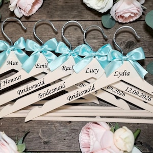 May include: Wooden hangers with silver hooks, personalized with names and roles like "Bride" and "Bridesmaid." Each hanger has a light blue satin bow. Some hangers include a date, "12.09.2026."