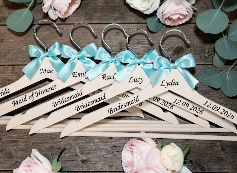 May include: Wooden hangers with silver hooks, personalized with names and roles like "Bride" and "Bridesmaid." Each hanger has a light blue satin bow. Some hangers include a date, "12.09.2026."